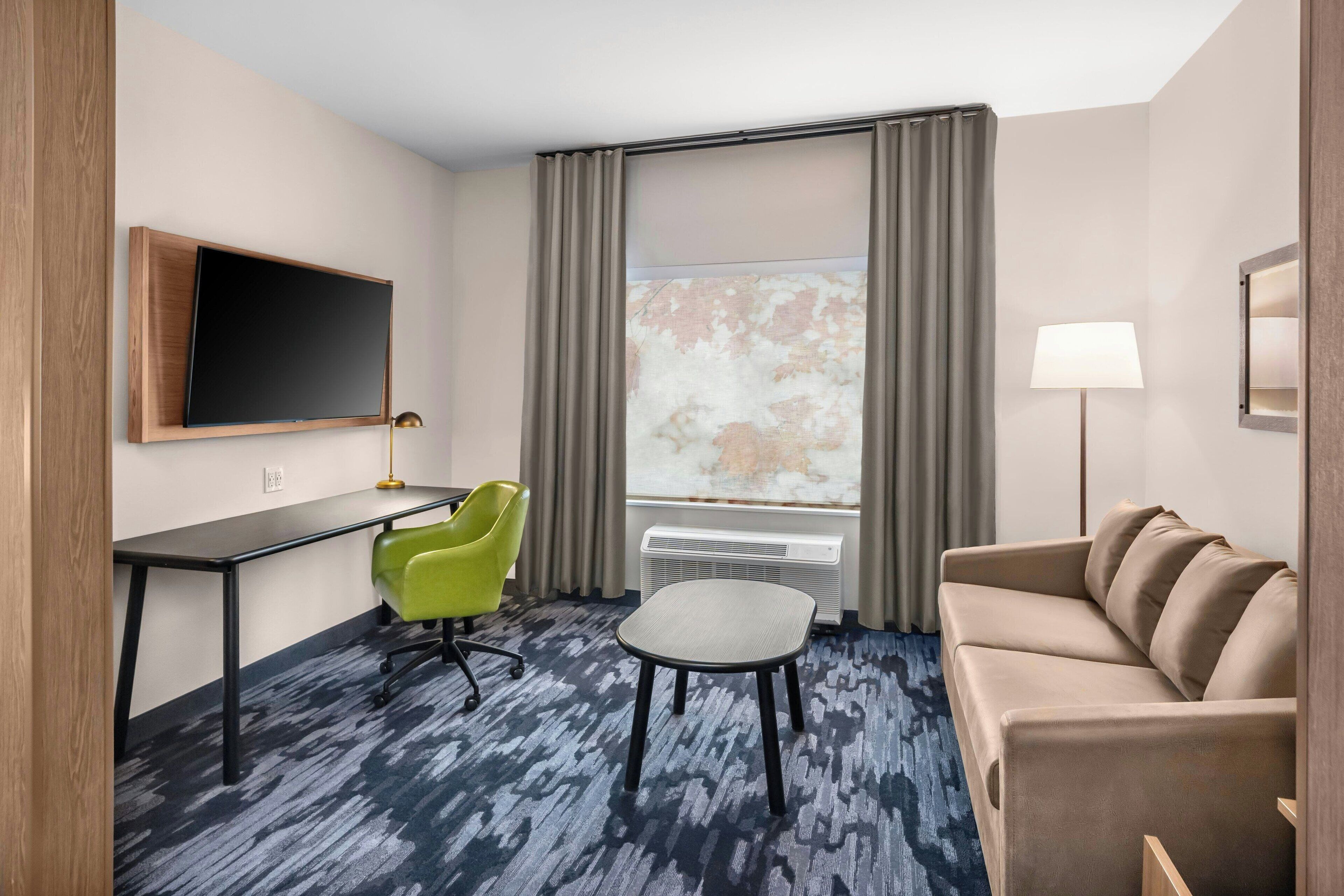 Habitación de hotel con escritorio, una silla verde, sofá, televisor montado en la pared, ventana con cortinas y una lámpara.
