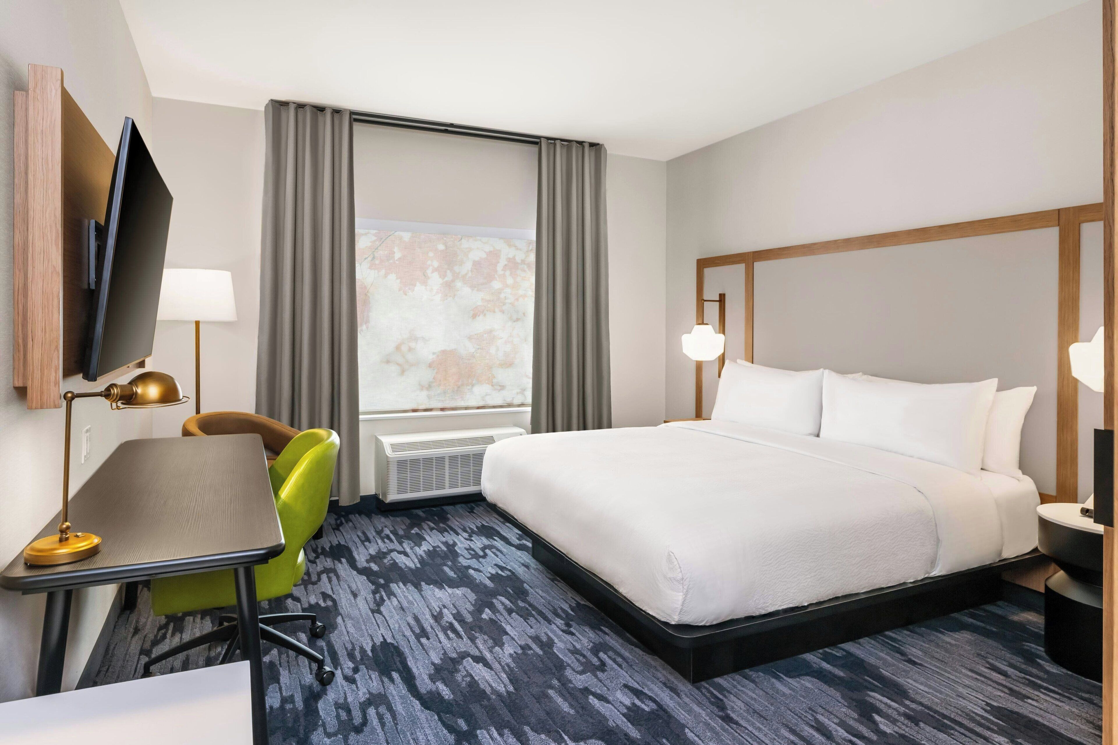 A hotel room with a bed, a desk with a lamp, a chair, a TV, and a window with curtains.