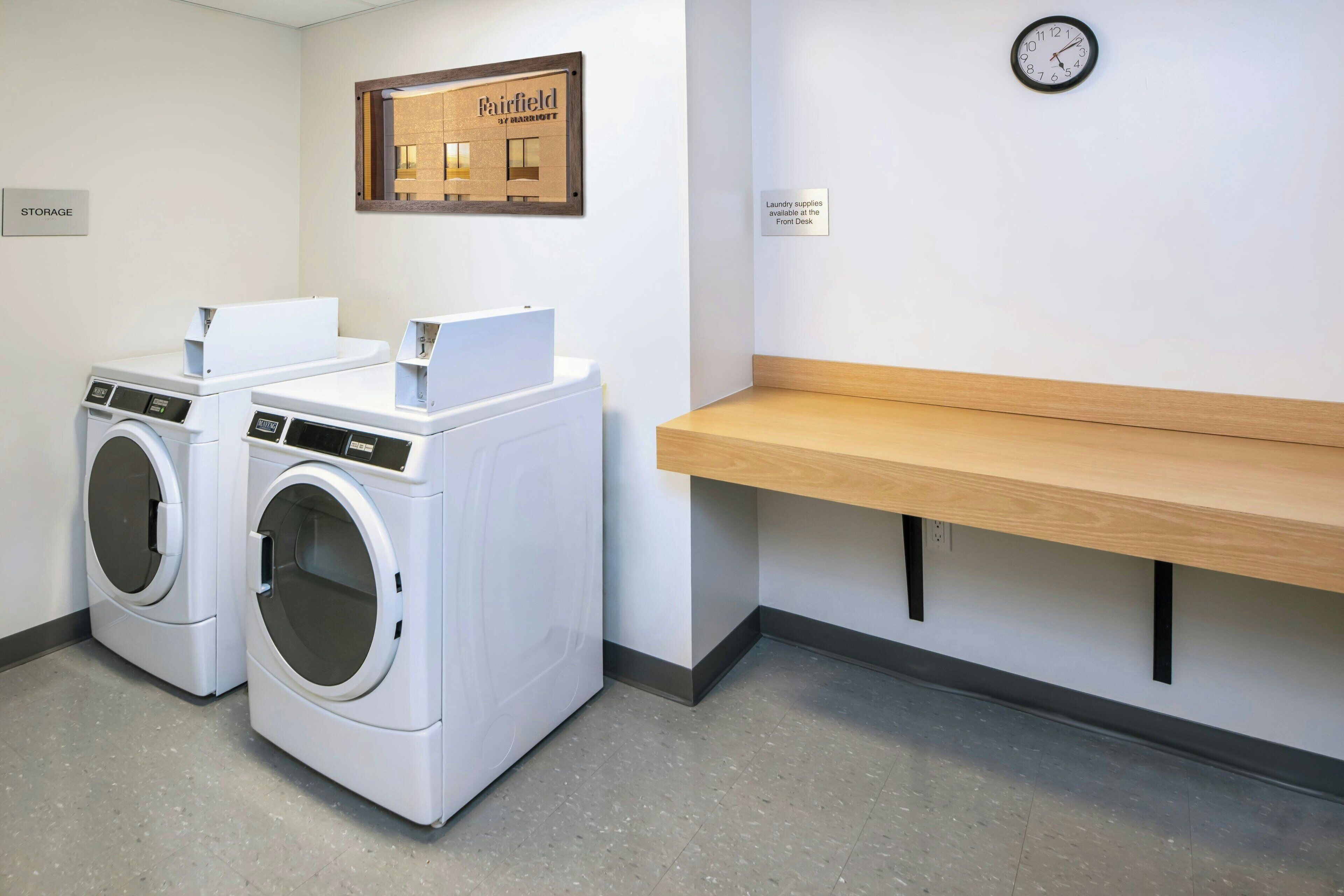 A laundry area with two front-loading washing machines, a wooden bench, a clock, and a sign indicating laundry supplies are available at the front desk.