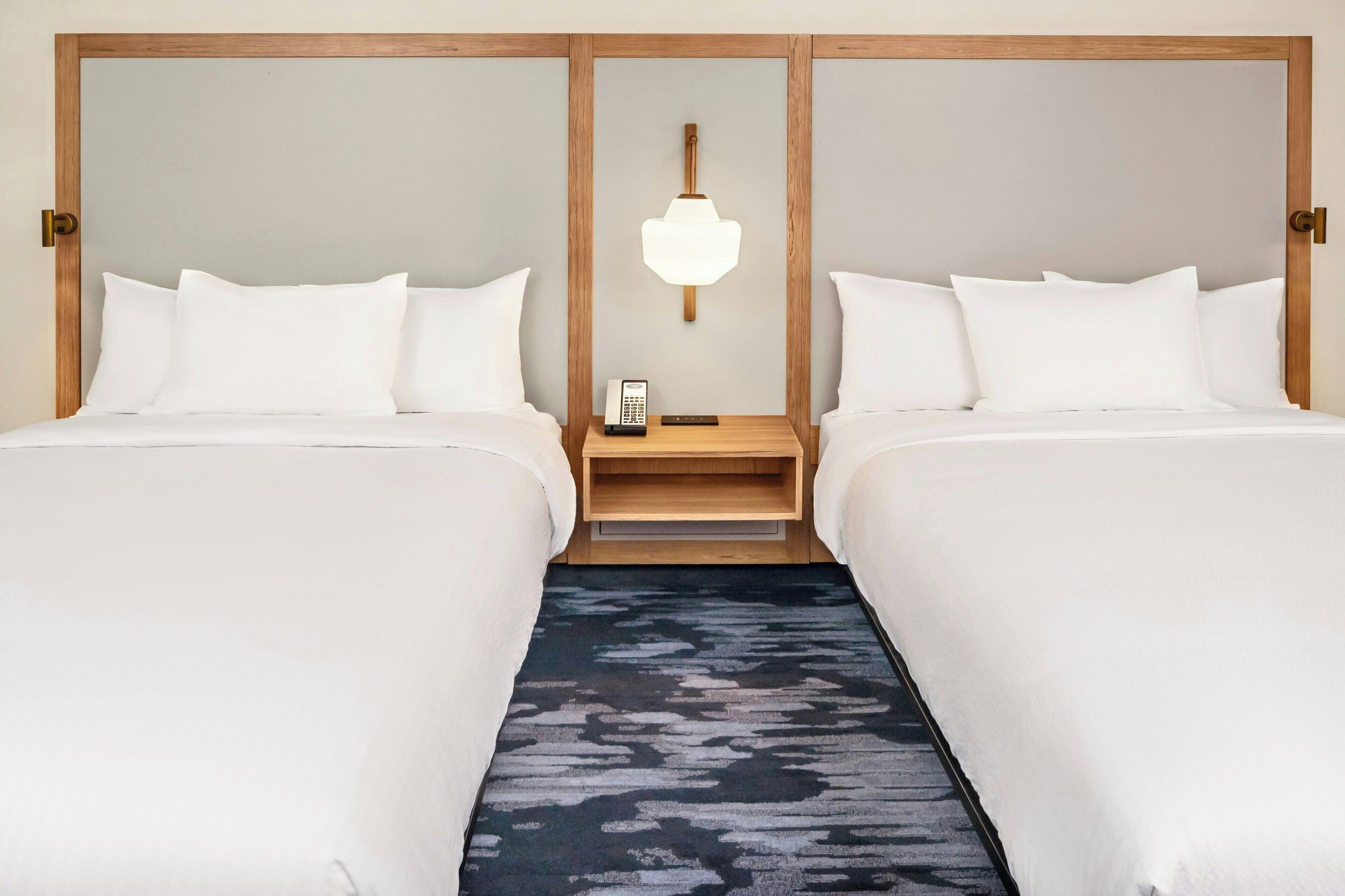 Une chambre d’hôtel avec deux lits, une tête de lit en bois, une table de chevet avec un téléphone et un tapis à motifs.