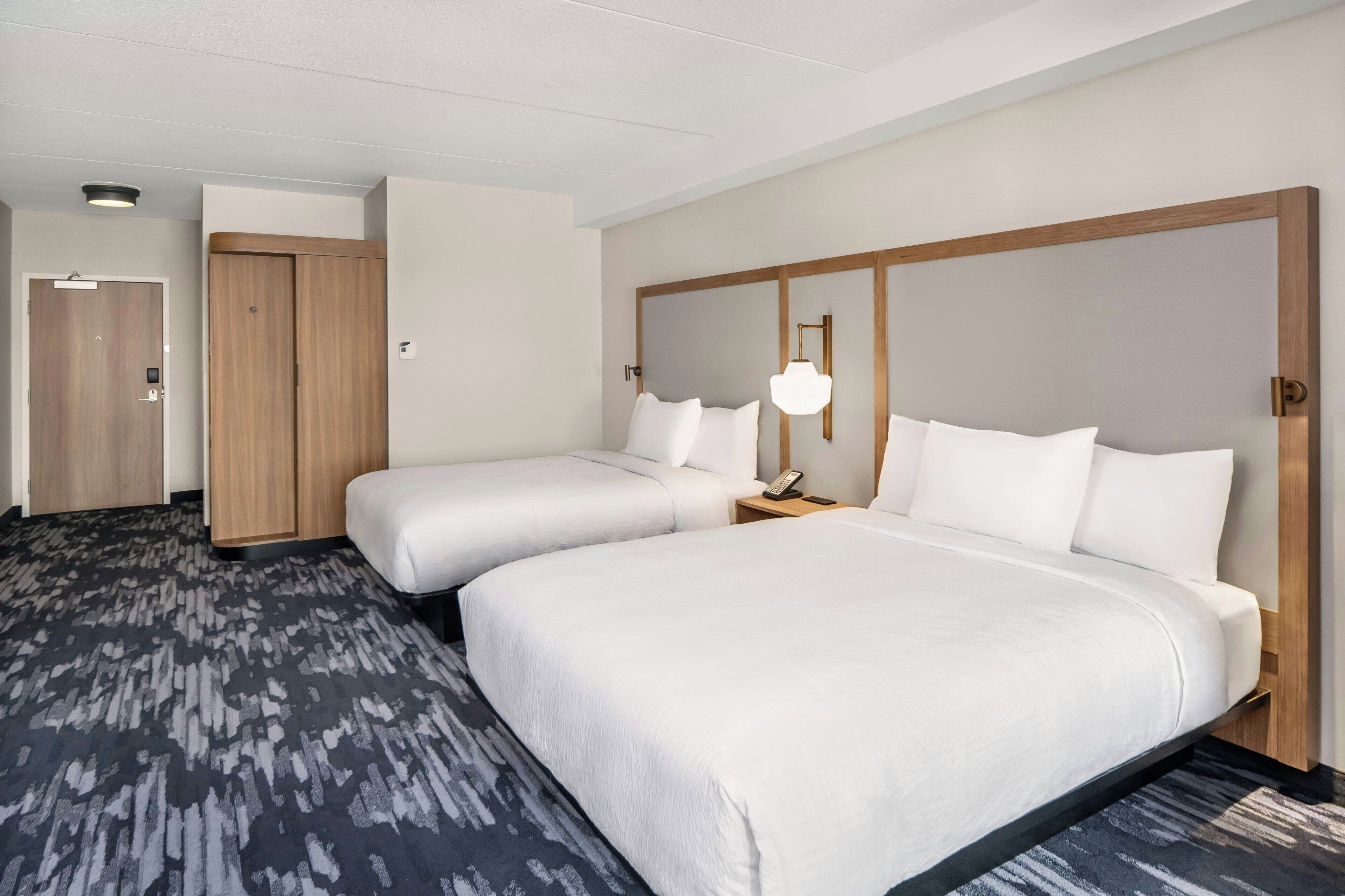 A hotel room with two beds, a wooden wardrobe, a desk with a lamp, and a carpeted floor.