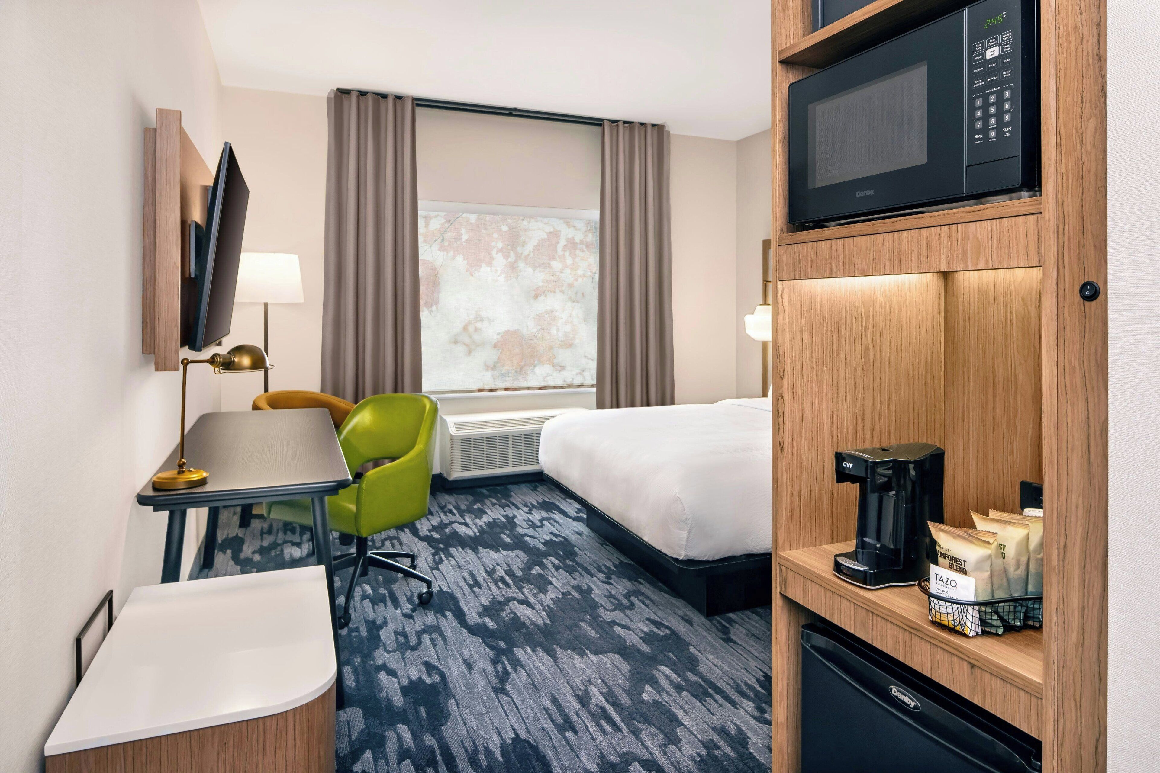 Quarto de hotel com cama, escrivaninha com luminária, cadeira, micro-ondas, cafeteira e estante com malas.