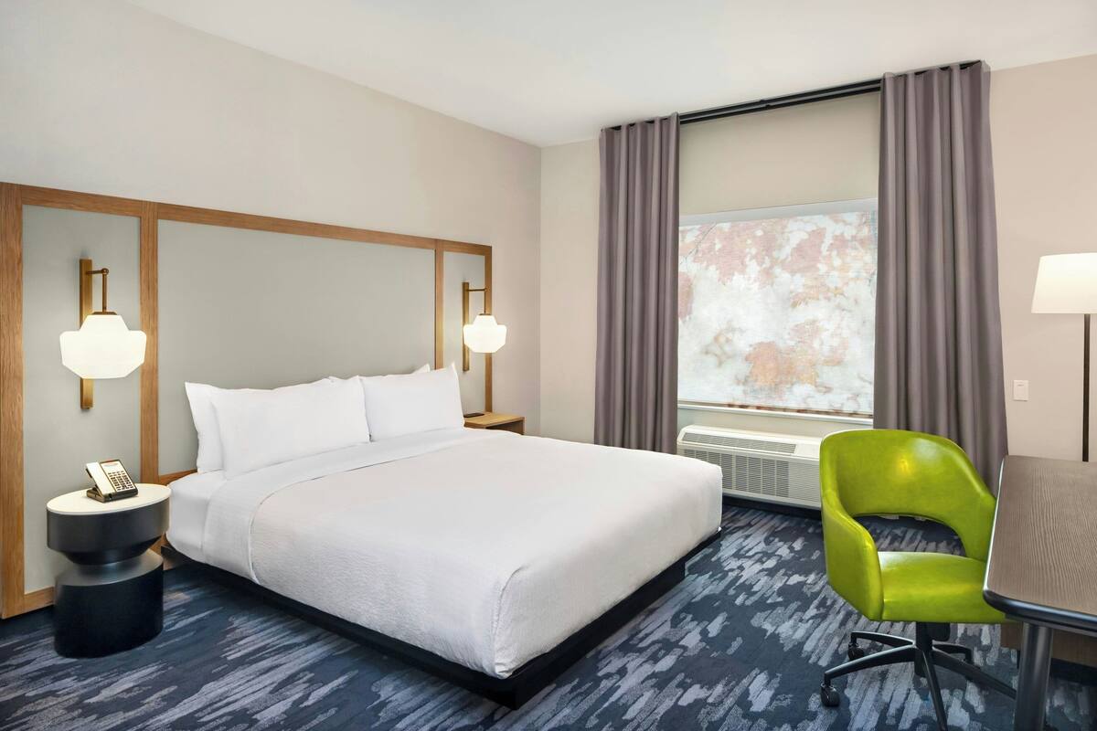 A hotel room with a large bed, a desk, a chair, a telephone, and a window with curtains.
