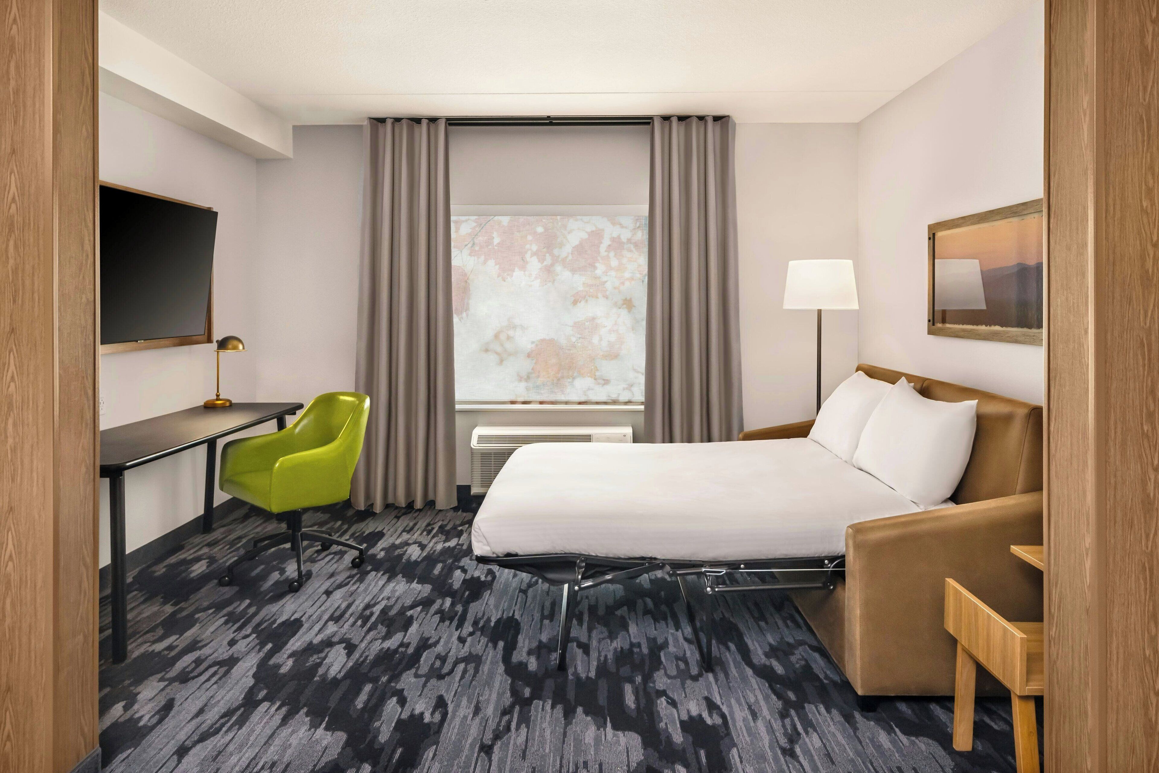 Una habitación de hotel con una cama, un escritorio, una silla verde, una lámpara y una ventana con cortinas.