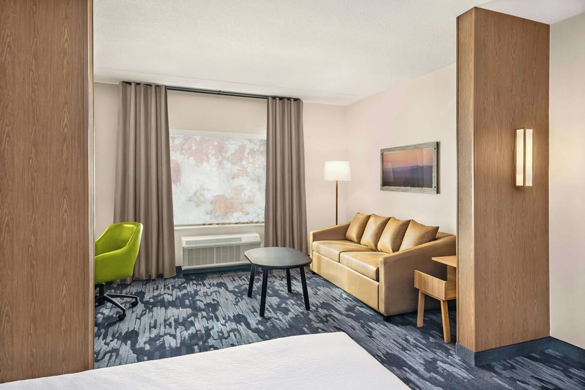 A hotel room with a bed, a sofa, a small table, a chair, a lamp, and a window with curtains.