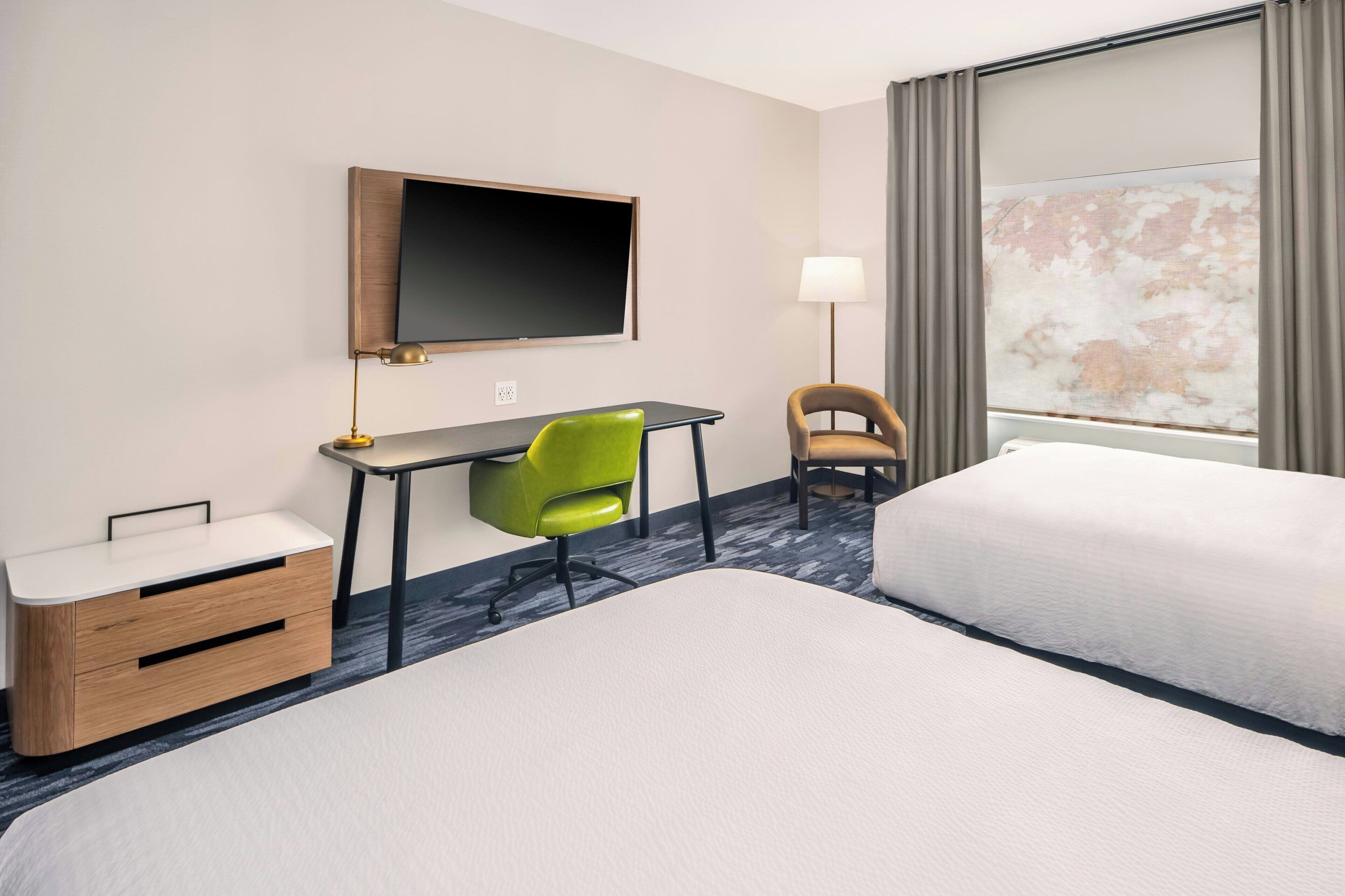 Une chambre d’hôtel avec deux lits, un bureau avec une chaise, une télévision, une lampe et une fenêtre avec des rideaux.