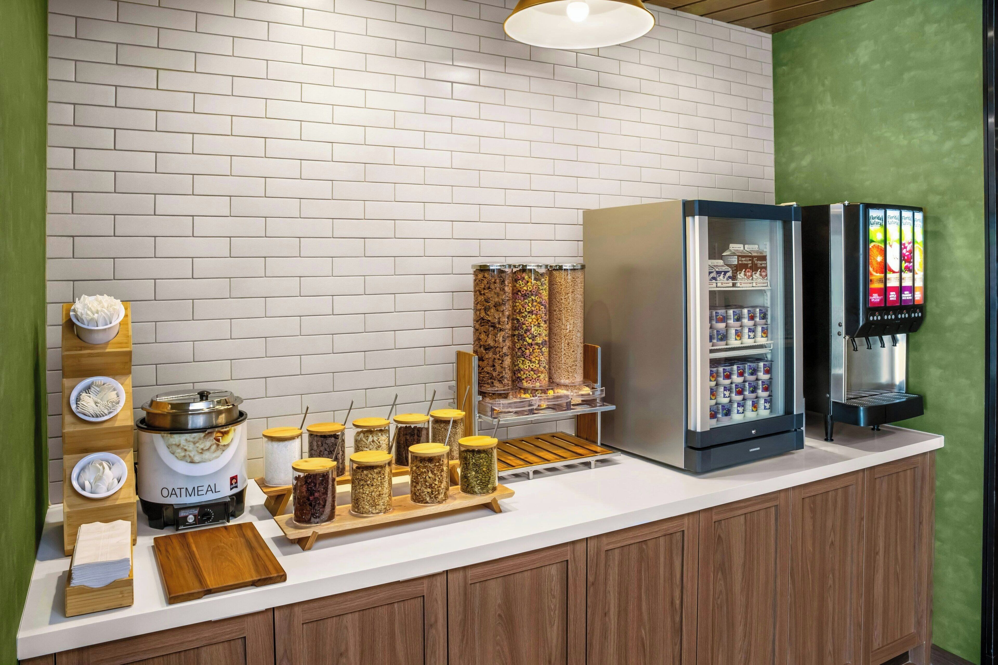Una barra de desayuno con dispensador de avena, frascos con conservas, un refrigerador expositor y un dispensador de bebidas.