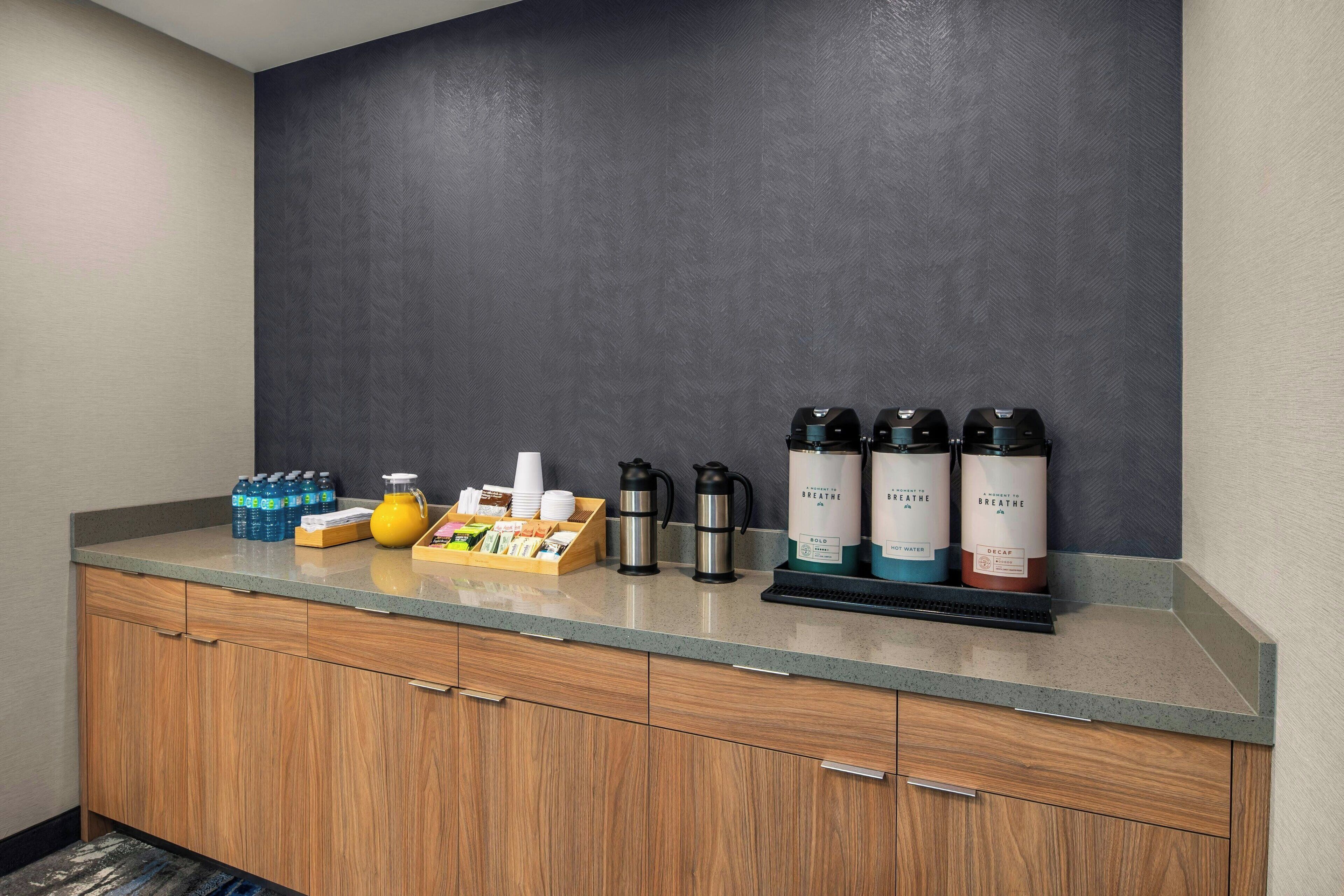 Una kitchenette con encimera gris, armarios de madera y diversos objetos como botellas, hervidor y cafetera.