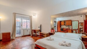A hotel room with a bed, a desk, a chair, a small table, a radiator, and a window with curtains.