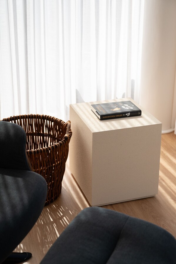 Una habitación moderna con una silla de mimbre, un sofá oscuro y una pequeña mesa blanca en cubo con un libro encima.