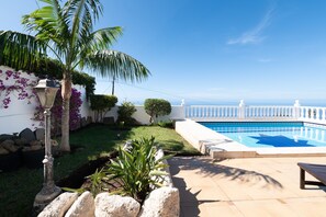 A pool area with a clear view of the sea, surrounded by a white fence and a well-maintained garden with a palm tree and purple flowers. - Charming Getaway in El Sauzal Enjoy Comfort Nature and a Private Pool (Sauzal)