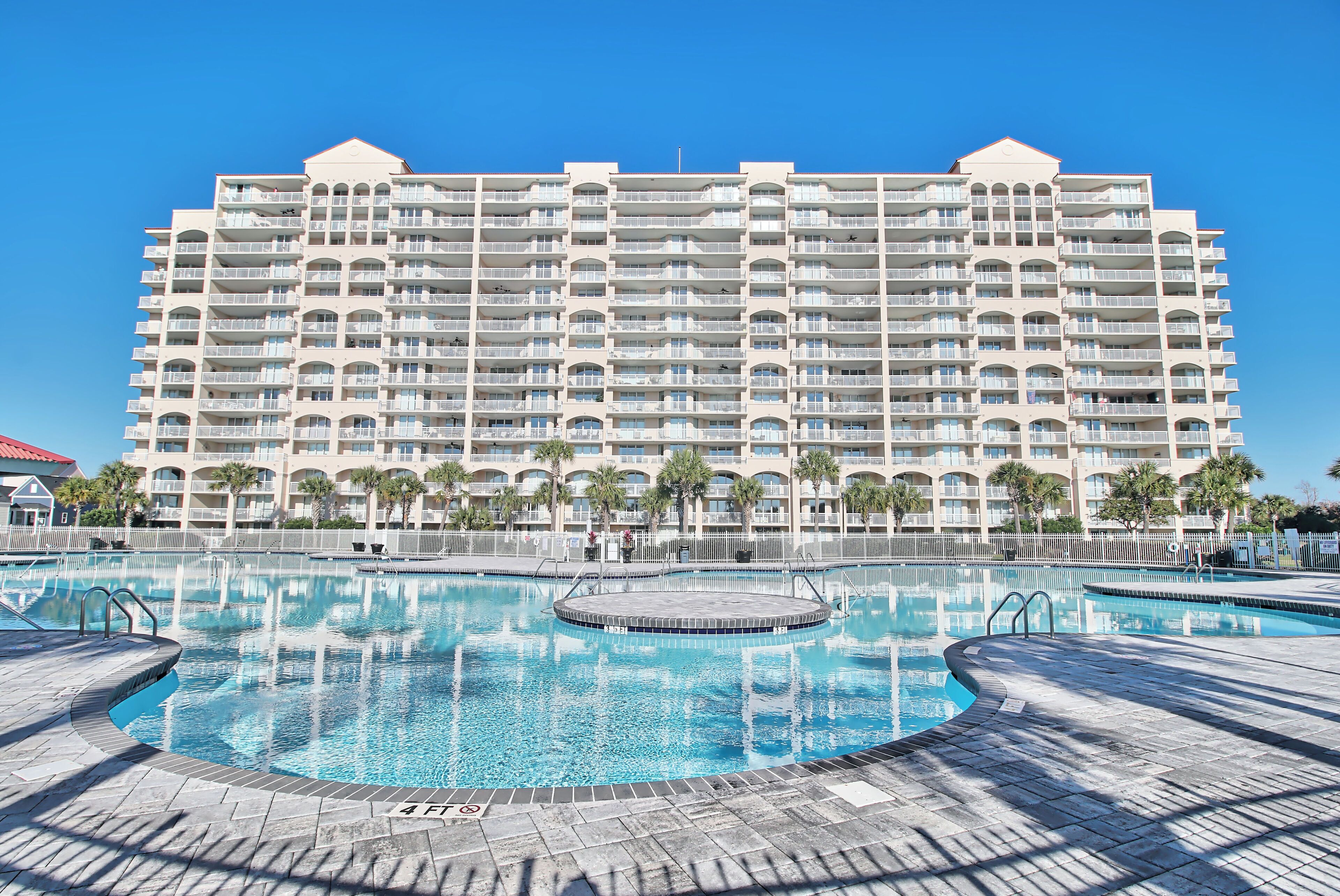 A large hotel with multiple balconies, a central swimming pool, and palm trees.