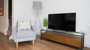 A living room with a grey sofa, a TV on a wooden stand, a floor lamp, and a staircase in the background.