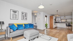 A modern living room with a grey sofa, ottoman, and a white rug. There is a kitchen area with wooden cabinets and a dining table.