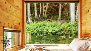 A wooden cabin bedroom with a bed, a desk, and a window overlooking a forest.