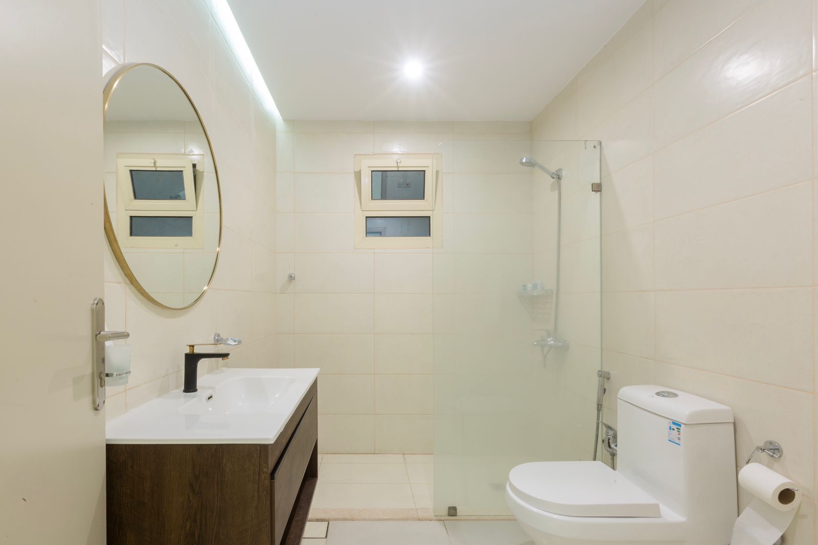 Een moderne badkamer met een witte wastafel, spiegel en toilet, een douchecabine van glas en een klein raam boven het toilet.