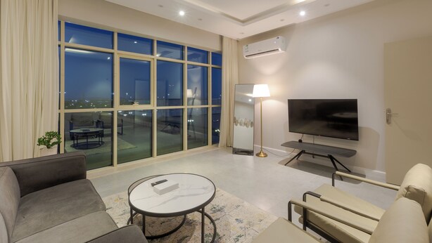 Una habitación de hotel moderna con un ventanal grande, un televisor de pantalla plana, un sofá, una mesa de centro y un pequeño balcón con mobiliario exterior.