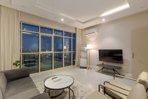 Una habitación de hotel moderna con un ventanal grande, un televisor de pantalla plana, un sofá, una mesa de centro y un pequeño balcón con mobiliario exterior.