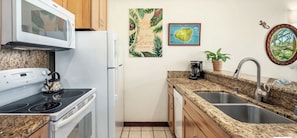 A compact kitchen with a stove, microwave, and sink. There are wooden cabinets, a coffee maker, and a potted plant.