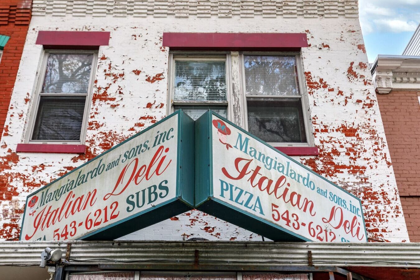 En bygning med afskallende hvid maling og røde detaljer. To skilte reklamerer for 'Mangialardo and sons, Inc. Italian Deli' og 'Mangialardo and sons, Inc. Pizza Subs'.