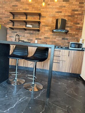 Kitchen area with a brick wall backdrop, featuring a shelf unit, range hood, microwave, and bar stool.