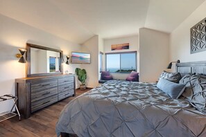 A bedroom with a large bed, a dresser, a mirror, a window with a view, and a plant.