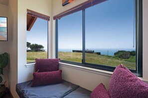 A room with a large window offering a view of the sea and grassy landscape. There are two purple cushions on a bench.