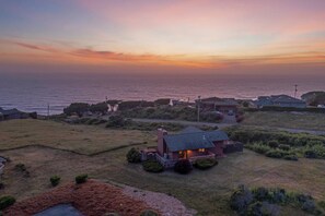 A coastal property with a prominent house featuring a dark roof and a visible chimney, surrounded by a well-maintained lawn and a small garden.