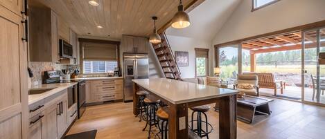 A modern kitchen with wooden cabinets, a central island with stools, a refrigerator, and a large window overlooking a deck.