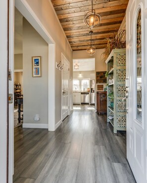 A hallway with wooden flooring, white walls, and a wooden ceiling. There are two doors, a shelf with decorative items, and a hanging light fixture.
