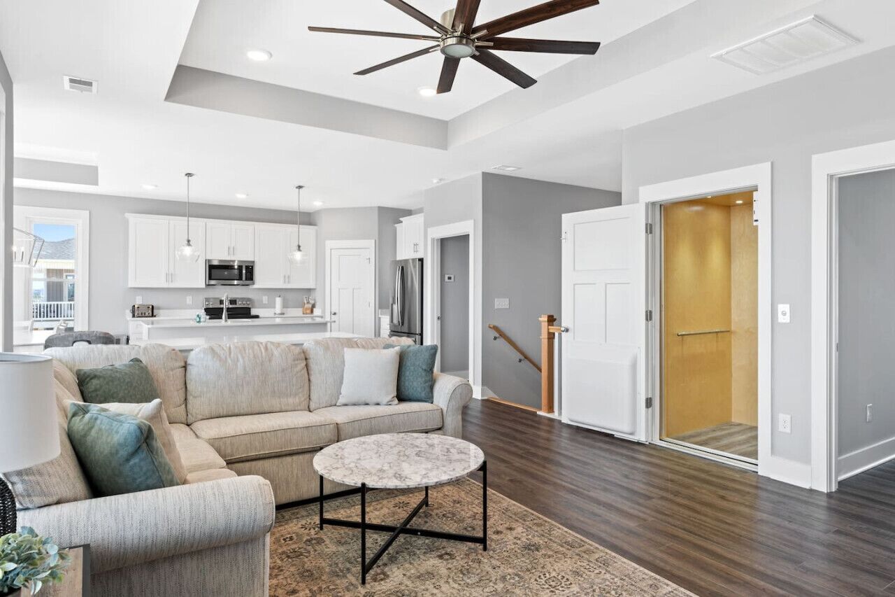 A spacious living room with a ceiling fan, a kitchen in the background, and a staircase leading to an upper level.