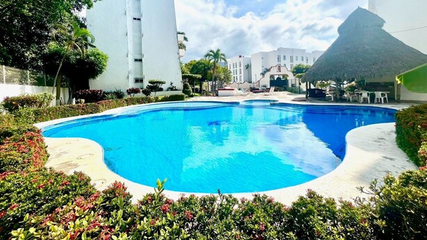 Una piscina rodeada de vegetación exuberante y edificios blancos, con una estructura de techo de paja distintiva al fondo.