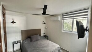 Un dormitorio con una cama, ventilador de techo, mesita de noche, lámpara, unidad de aire acondicionado y televisor montado en la pared.