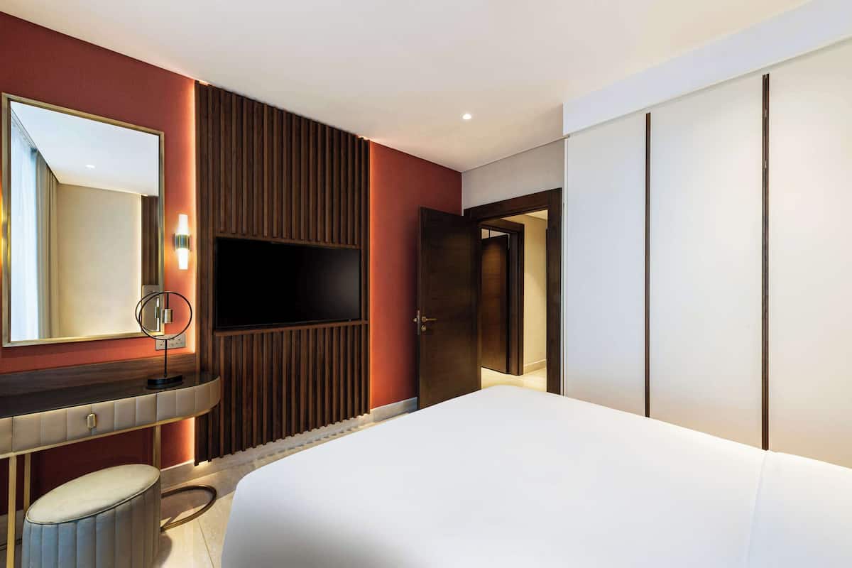 A modern hotel room with a large bed, a vanity with a mirror, a television, and a wardrobe.