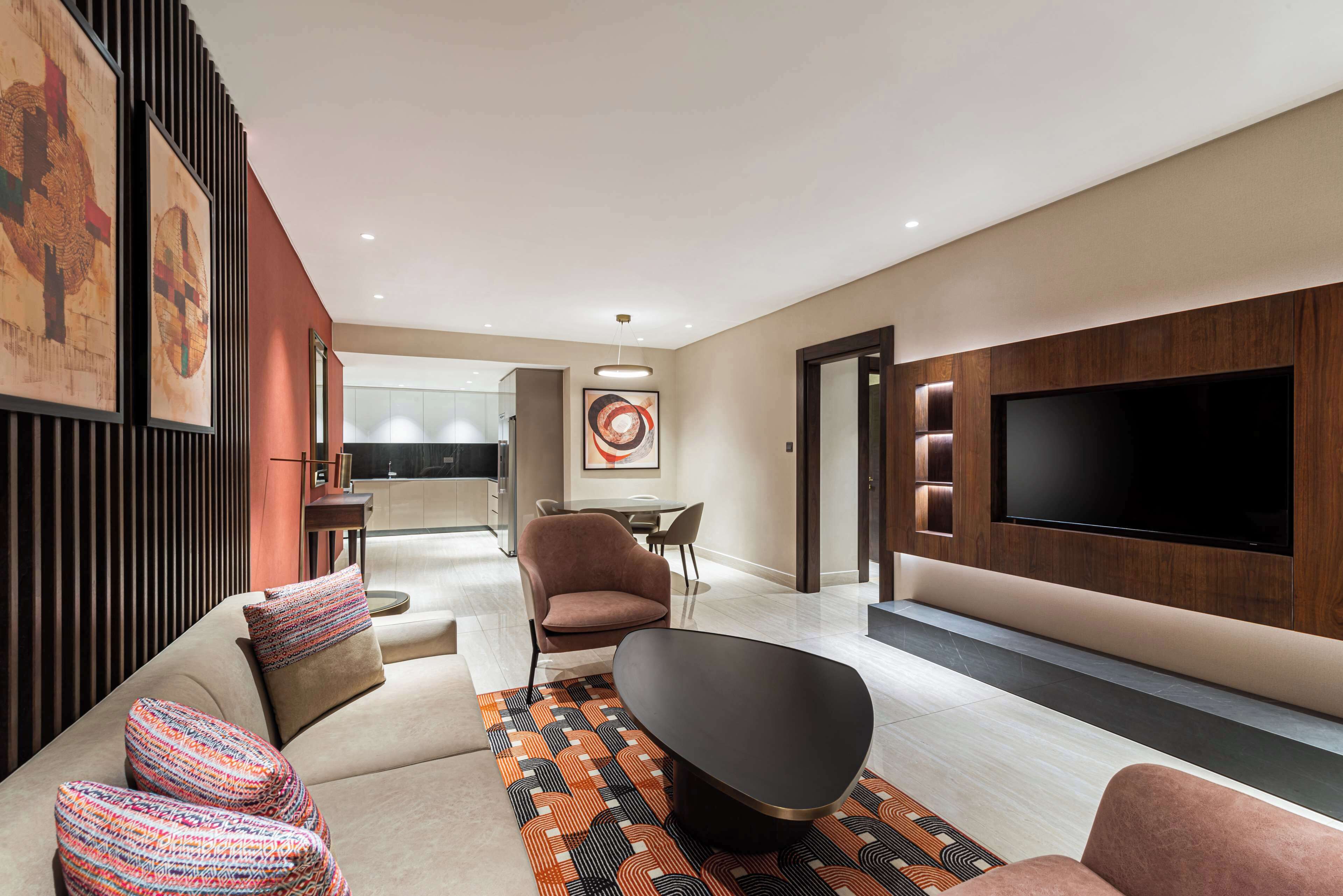 Ein modernes Wohnzimmer mit einer Couch, Sesseln, einem Couchtisch und einem flachbildfernseher an der Wand. Im Hintergrund befindet sich ein Essbereich mit Tisch und Stühlen.