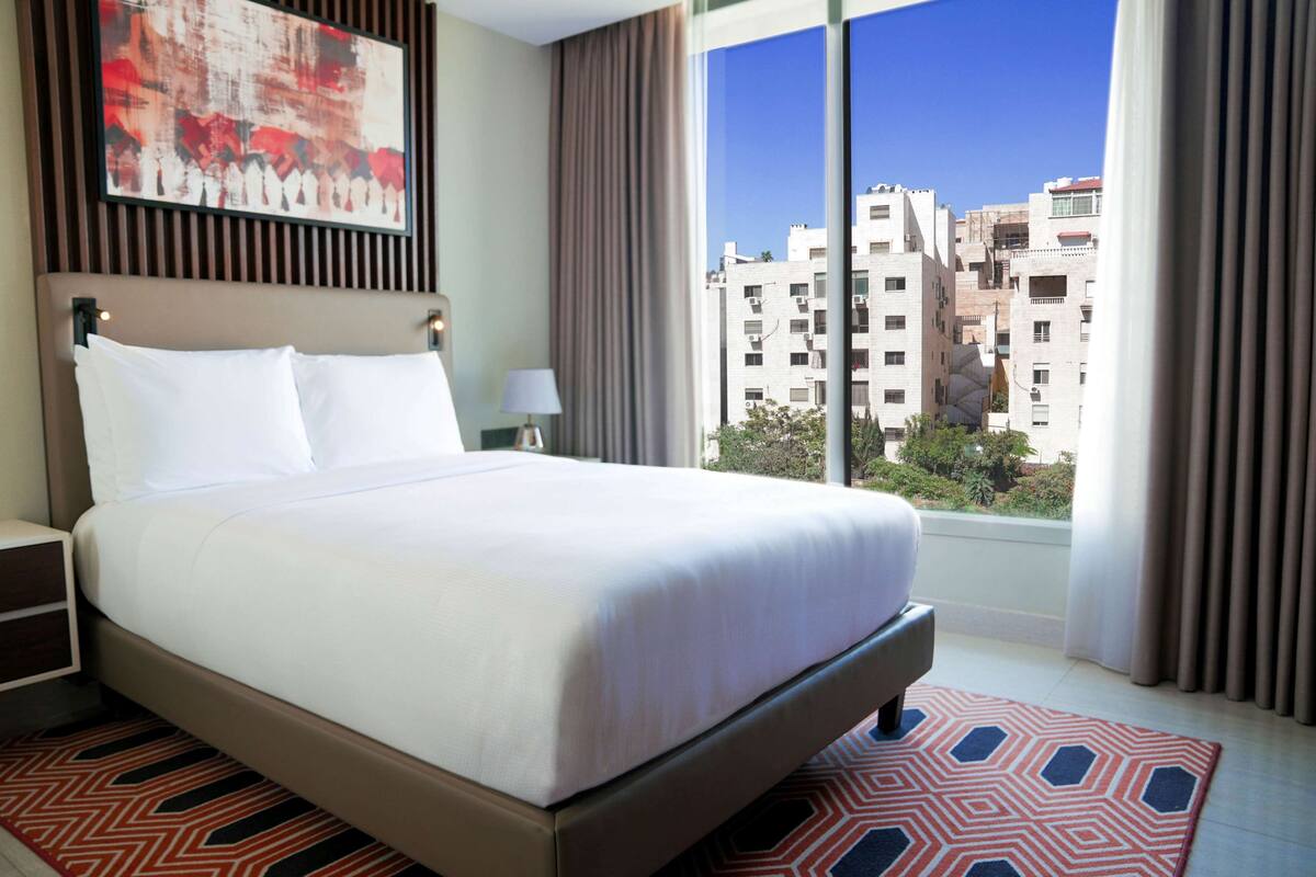 A hotel room with a large bed, a nightstand, a lamp, and a view of the cityscape through large windows.