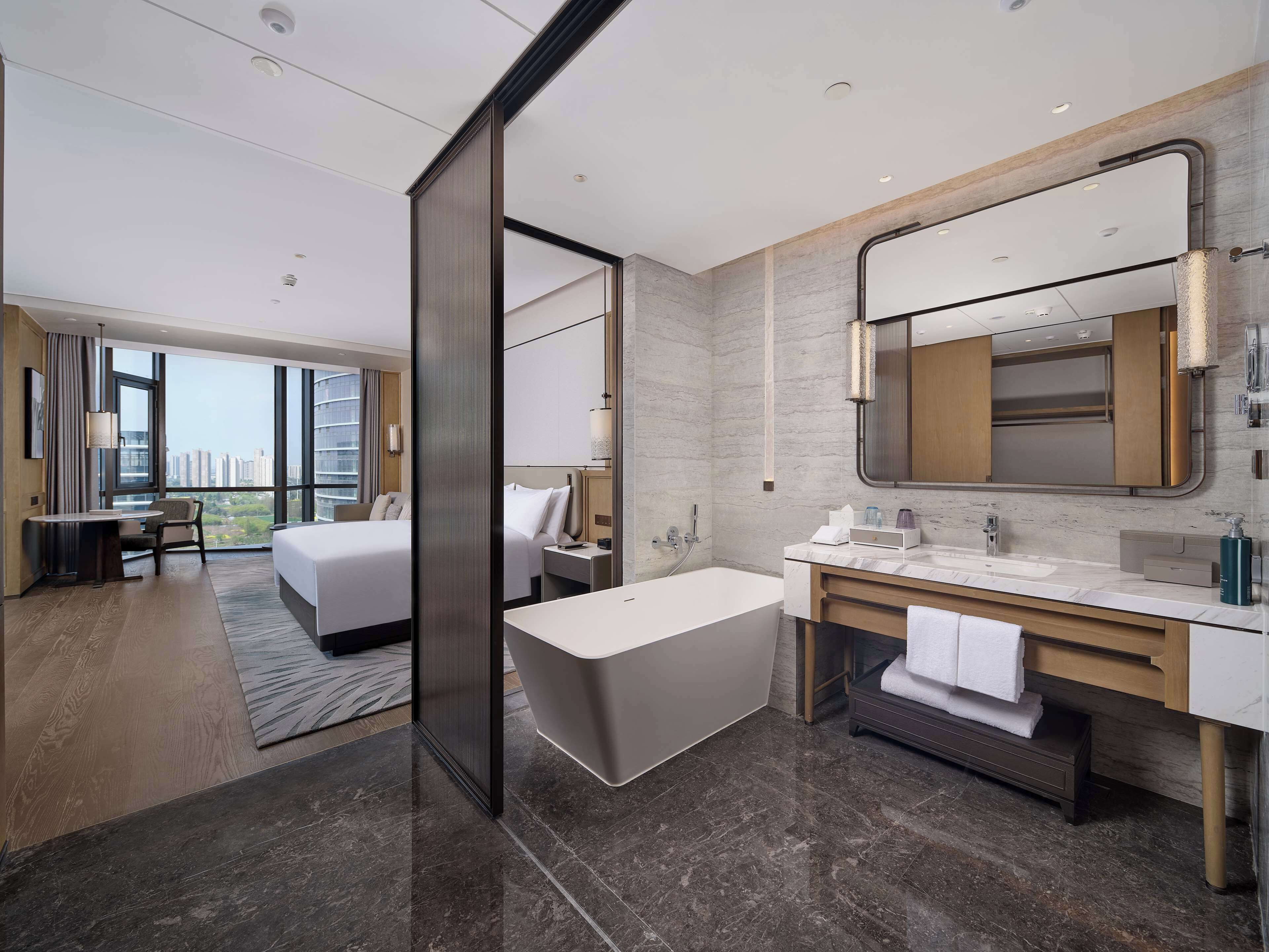 Une chambre d’hôtel moderne dotée d’un grand lit, d’un coin salon séparé, d’une baignoire et d’une salle de bain avec un grand miroir et un lavabo.