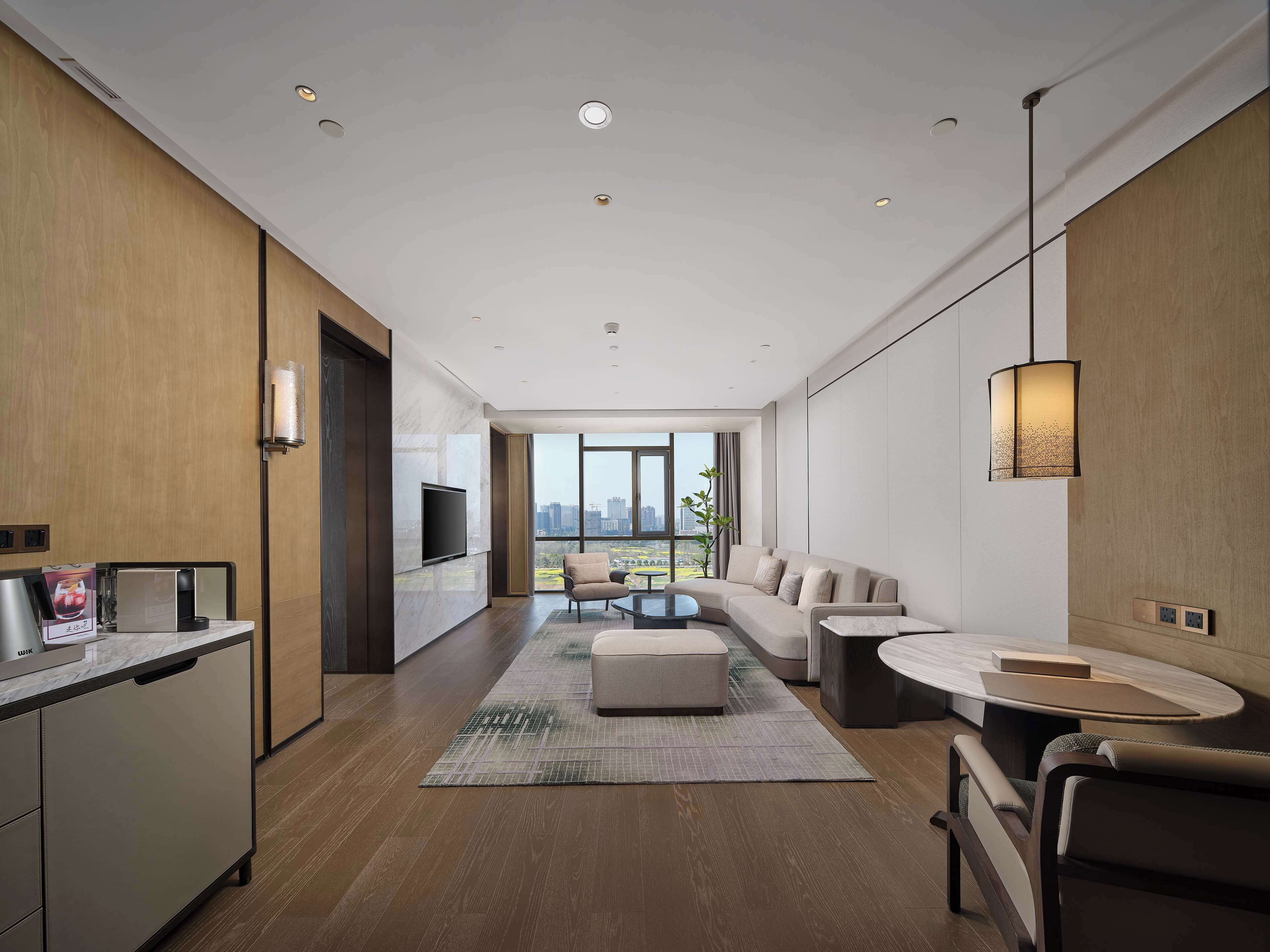 Una sala de estar moderna con un sofá, una mesa de centro y un televisor montado en la pared. Un ventanal grande ofrece vistas de la ciudad.