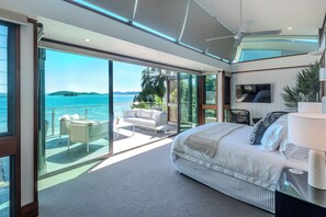 A modern bedroom with a large bed, a TV, and a balcony with a view of the sea.