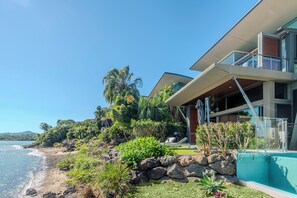A modern beachfront property with a pool, lush vegetation, and a view of the ocean.