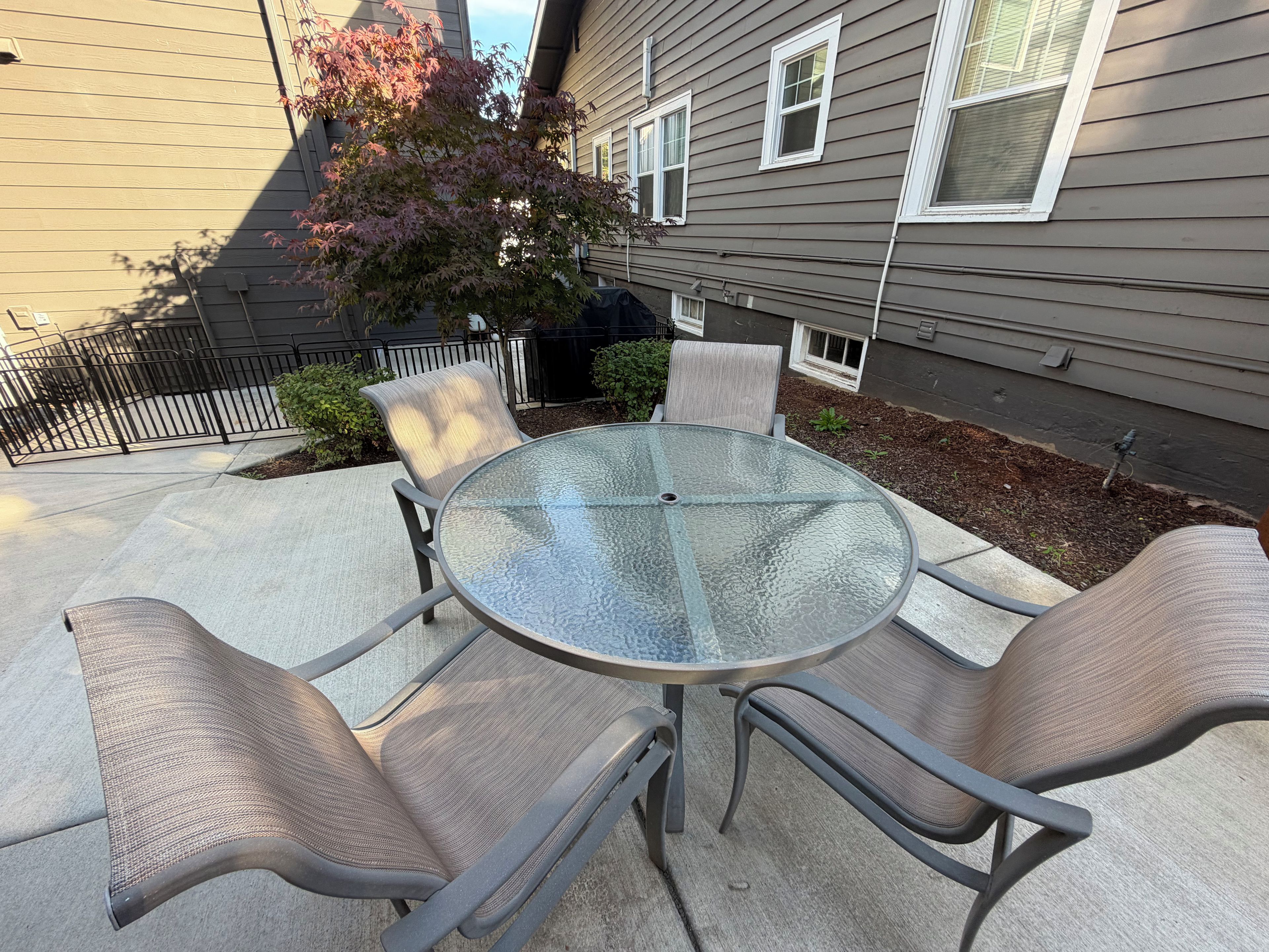 A patio with a glass table and four chairs, a small tree, and a house in the background.