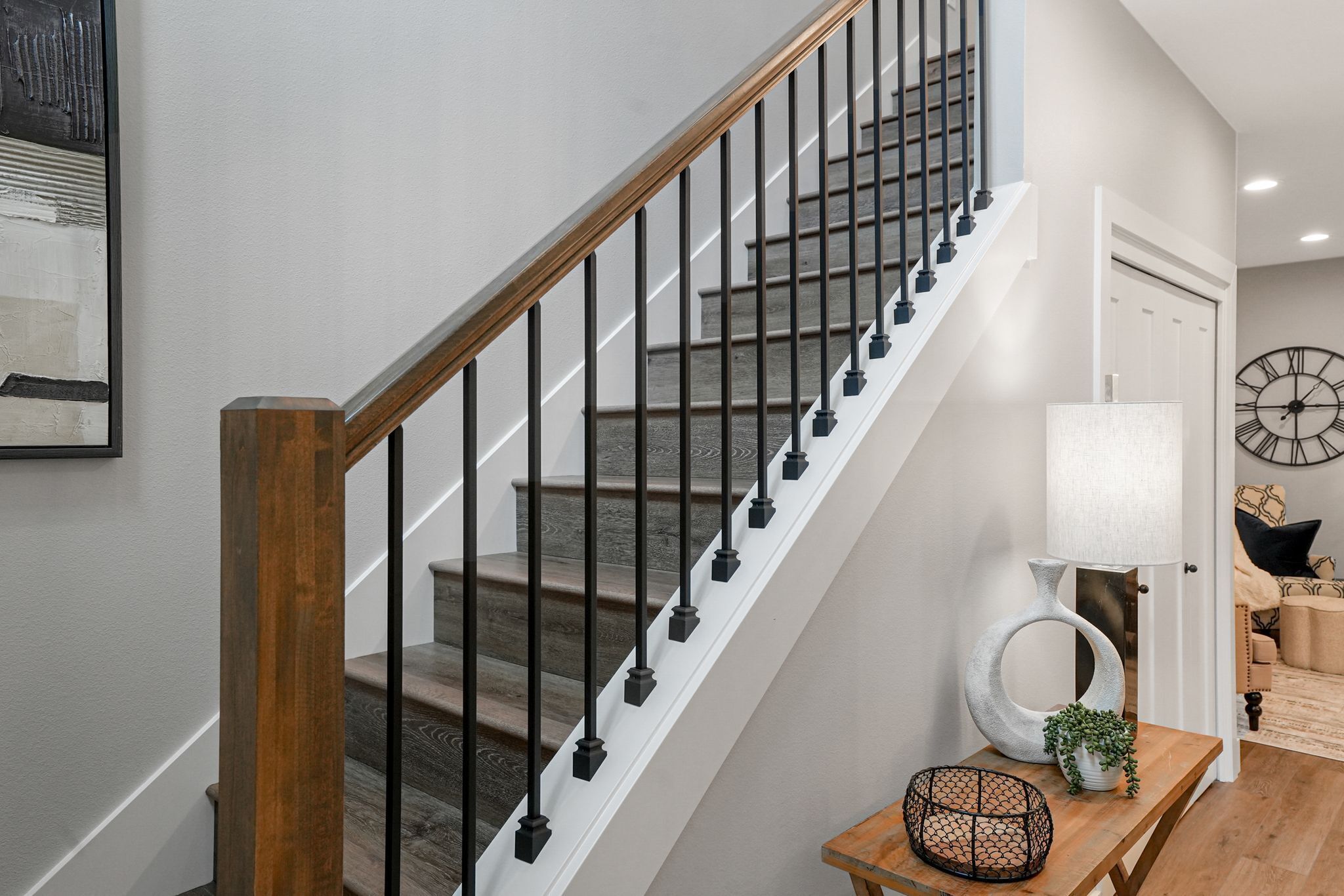 Una escalera con barandillas de madera y balaustres negros, que conduce a una habitación con una puerta blanca, una lámpara y un reloj en la pared.