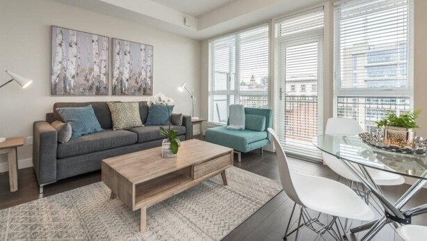 Una sala de estar moderna con un sofá gris, un sillón azul turquesa y una mesa de comedor de vidrio con sillas blancas. Hay pinturas abstractas en la pared y un amplio ventanal con persianas.