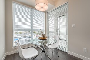 A dining area with a glass table, two white chairs, and a view of buildings through large windows with blinds. - Bright Old Town Suite with City Views (Victoria Central British Columbia Provincial Government)