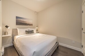 A bedroom with a large bed, two bedside tables, a nightstand, a wall-mounted painting, and a wooden floor. - Bright Old Town Suite with City Views (Victoria Central British Columbia Provincial Government)