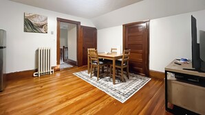 Un coin repas avec une table et des chaises en bois, un radiateur, un téléviseur posé sur un meuble et un tapis à motifs.