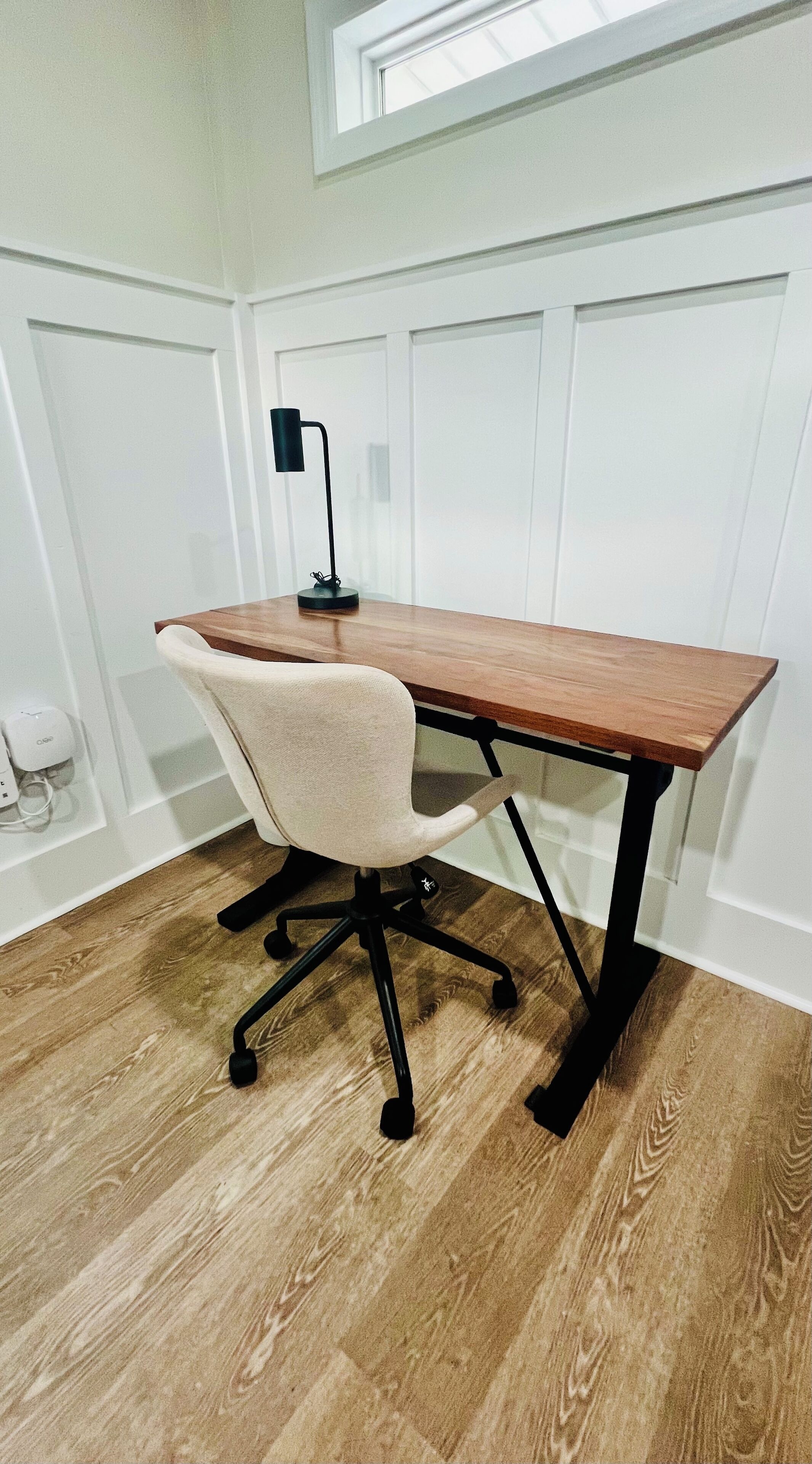 Een houten bureau met een zwart metalen frame, een witte bureaustoel en een zwarte bureaulamp.
