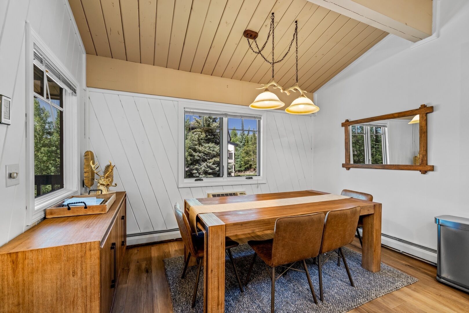 A dining area with a wooden table and chairs, a chandelier, and a view of trees through a window.