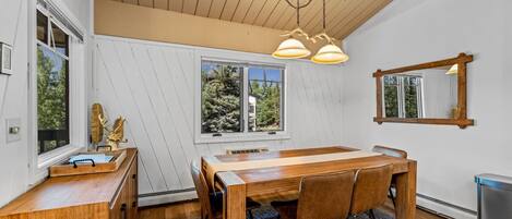 A dining area with a wooden table and chairs, a chandelier, and a view of trees through a window.