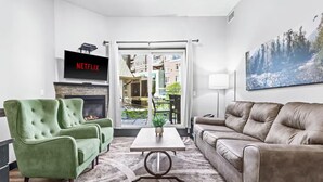 A modern living room with a green armchair, a brown sofa, a fireplace, a flat-screen TV, and a large window with a view of buildings. - Mountain Getaway in Canmore! Sleeps 6 - Pool & Hottub (Canmore)